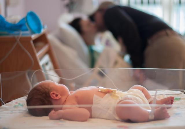 Savannah and Eric Rheude share a kiss in the background, as their newborn baby girl Ella June rests in her bassinet at the Women's Pavilion at OSF Saint Anthony's Health Center.