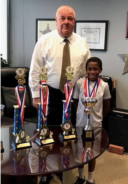 Madison County Sheriff John D. Lakin and tumbling/trampoline star Micah Miner of Glen Carbon with some of Micah's trophies and medals.